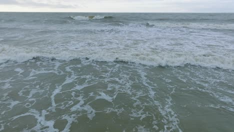 Aerial-view-of-big-stormy-waves-breaking-against-the-white-sand-beach,-overcast-day,-seashore-dunes-damaged-by-waves,-coastal-erosion,-climate-changes,-wide-low-drone-shot-moving-forward