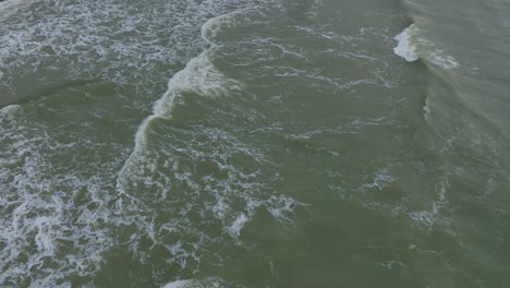 Aerial-birdseye-view-of-big-stormy-waves-breaking-against-the-white-sand-beach,-overcast-day,-seashore-dunes-damaged-by-waves,-coastal-erosion,-climate-changes,-wide-drone-shot-moving-forward