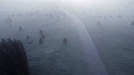 Aerial-drone-shot-over-a-foggy-frozen-Thetford-cemetery-on-London-Road-showing-snow-filled-grave-yard-on-both-sides-of-a-path