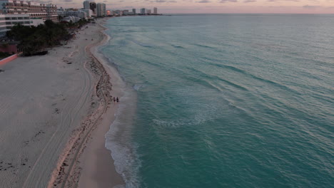 Drone-view-following-couple-walking-on-white-sand-beach-at-dusk,-Cancun,-Mexico