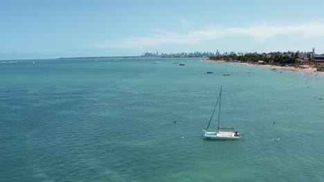 Dolly-in-tilt-up-aerial-shot-of-Bessa-beach-in-the-tropical-capital-city-of-Joao-Pessoa,-Paraiba,-Brazil-passing-a-sail-boat-with-people-enjoying-the-ocean-and-skyscrapers-in-the-background