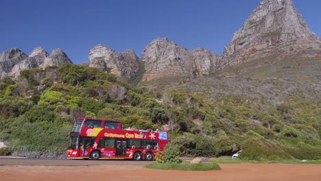 Red-double-decker-tour-bus-drives-past-steep-rugged-Cape-town-mountains