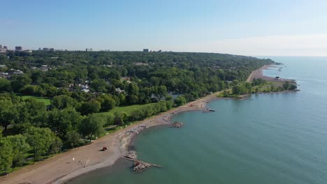Aerial-shot-flying-along-the-coastline-of-Lake-Ontario,-with-a-beach-and-a-tennis-court-in-the-summer