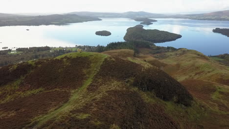 Aerial-orbits-man-on-hill-overlooking-Scottish-loch-on-cloudy-day
