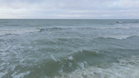 Aerial-establishing-view-of-big-stormy-waves-breaking-against-the-white-sand-beach,-overcast-day,-seashore-dunes-damaged-by-waves,-coastal-erosion,-climate-changes,-wide-low-drone-shot-moving-forward