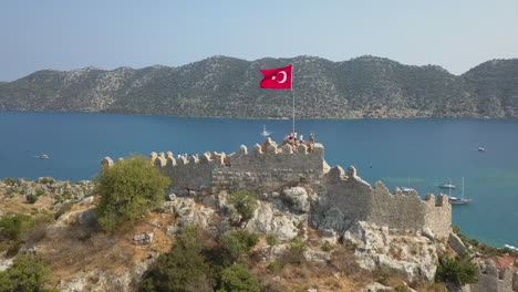 Aerial-orbits-Simena-Castle-ruin-on-rock-hilltop-flying-Flag-of-Turkey