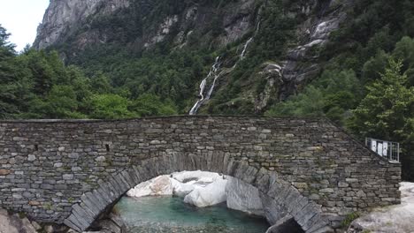 Mountain-river-streaming-underneath-old-cobblestone-bridge-in-Switzerland,-ascend-view
