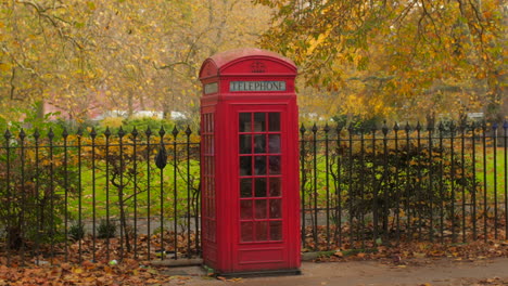 Vintage-Red-Phone-Booth-On-The-Urban-Park-With-Metal-Fence-During-Fall-Season-In-London,-UK