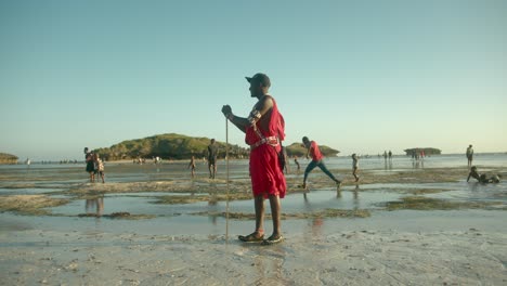 Guerrero-Masai-De-Pie-En-La-Playa-Con-Lugareños-Caminando-En-Segundo-Plano-En-Un-Día-Ventoso-En-Watamu,-Kenia