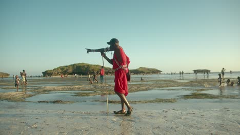 Guerrero-Masai-Con-Ropa-Tradicional-Roja-Parado-En-La-Playa-Sosteniendo-Un-Poste-De-Madera-Y-Apuntando-En-Dirección-A-Watamu,-Kenia