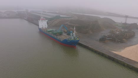 Aerial-establishing-view-of-wood-terminal-crane-loading-timber-into-the-cargo-ship,-Port-of-Liepaja-,-lumber-log-export,-overcast-day-with-fog-and-mist,-drone-moving-forward,-tilt-down