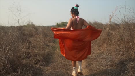 The-back-of-a-carefree-attractive-Asian-female-embracing-the-freedom-of-nature,-her-oversized-shirt-fluttering-in-the-morning-breeze-as-she-walks-along-a-path-in-a-field,-India