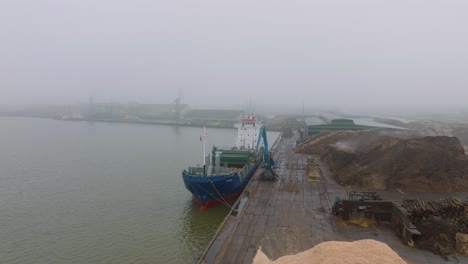 Aerial-establishing-view-of-wood-terminal-crane-loading-timber-into-the-cargo-ship,-Port-of-Liepaja-,-lumber-log-export,-overcast-day-with-fog-and-mist,-wide-drone-shot-moving-forward-slow