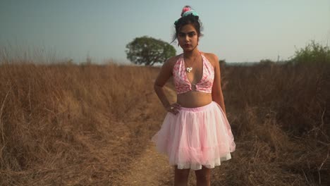 An-attractive-Asian-female-wearing-a-pink-tutu-dress,-pushing-her-hair-back-over-her-ears-as-she-stands-in-a-breezy-dry-grass-field-surrounded-by-nature,-India