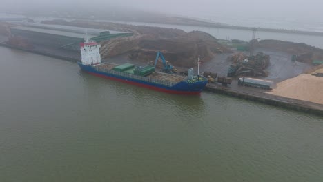 Aerial-establishing-view-of-wood-terminal-crane-loading-timber-into-the-cargo-ship,-Port-of-Liepaja-,-lumber-log-export,-overcast-day-with-fog-and-mist,-drone-shot-moving-forward,-tilt-down