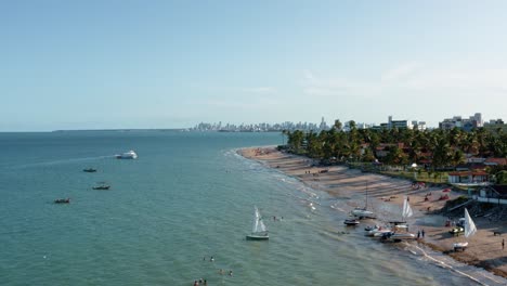 Tilt-up-aerial-wide-shot-of-the-tropical-Bessa-beach-in-the-capital-city-of-Joao-Pessoa,-Paraiba,-Brazil-with-people-enjoying-the-ocean,-small-fishings-boats-and-skyscrapers-in-the-background