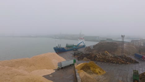 Aerial-establishing-view-of-wood-terminal-crane-loading-timber-into-the-cargo-ship,-Port-of-Liepaja-,-lumber-log-export,-day-with-fog-and-mist,-wood-chip-piles,-wide-drone-shot-moving-forward