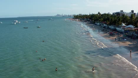 Dolly-in-tilt-up-aerial-wide-shot-of-the-tropical-Bessa-beach-in-the-capital-city-of-Joao-Pessoa,-Paraiba,-Brazil-with-people-enjoying-the-ocean,-small-fishings-boats-and-skyscrapers-in-the-background