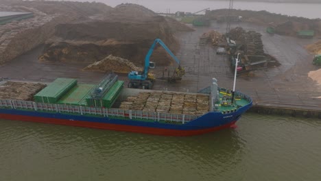 Aerial-establishing-view-of-wood-terminal-crane-loading-timber-into-the-cargo-ship,-Port-of-Liepaja-,-lumber-log-export,-overcast-day-with-fog-and-mist,-drone-shot-moving-forward,-tilt-down