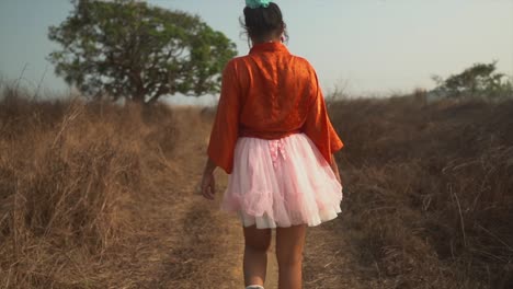 A-slow-motion-shot-of-the-back-of-an-outgoing-carefree-Asian-female-wearing-a-pink-tutu-dress,-stretching-her-arms-out-while-walking-along-a-pathway-in-a-dry-grass-field,-India