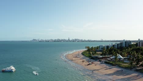 Aerial-drone-wide-shot-of-the-tropical-Bessa-beach-in-the-capital-city-of-Joao-Pessoa,-Paraiba,-Brazil-with-people-enjoying-the-ocean,-a-small-tour-boat,-jet-ski,-and-skyscrapers-in-the-background