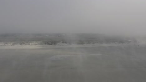 Drone-flying-above-very-foggy-Beach-along-the-wind-and-the-swirled-up-sand-towards-dunes