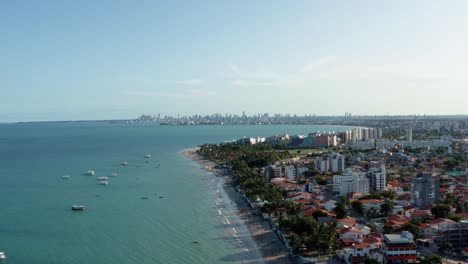 Descending-aerial-drone-extreme-wide-shot-of-the-tropical-Bessa-beach-in-the-city-of-Joao-Pessoa,-Paraiba,-Brazil-with-people-enjoying-the-ocean,-tour-boats,-and-large-cityscape-in-the-background