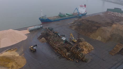 Aerial-establishing-view-of-wood-terminal-crane-loading-timber-into-the-cargo-ship,-Port-of-Liepaja-,-lumber-log-export,-overcast-day-with-fog-and-mist,-birdseye-drone-shot-moving-forward