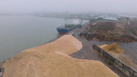 Aerial-establishing-view-of-wood-terminal-crane-loading-timber-into-the-cargo-ship,-Port-of-Liepaja-,-lumber-log-export,-overcast-day-with-fog-and-mist,-wood-chip-piles,-wide-drone-moving-back