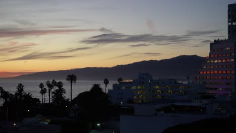 Santa-Monica-Coastline-during-sunset,-Palm-trees-silhouette-along-the-shoreline,-Pan-shot