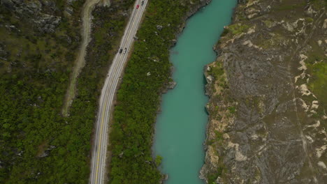 Aerial-top-down-view-of-a-forest-road-and-where-vehicles-pass-next-to-a-turquoise-river-in-a-beautiful-valley