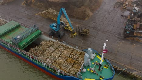 Aerial-establishing-view-of-wood-terminal-crane-loading-timber-into-the-cargo-ship,-Port-of-Liepaja-,-lumber-log-export,-overcast-day-with-fog-and-mist,-wide-birdseye-drone-shot