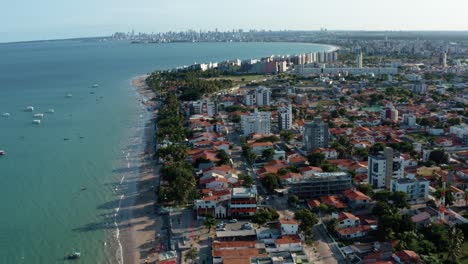 Tilt-up-rising-aerial-drone-extreme-wide-shot-of-the-tropical-Bessa-beach-in-the-city-of-Joao-Pessoa,-Paraiba,-Brazil-with-people-enjoying-the-ocean,-tour-boats,-and-large-cityscape-in-the-background