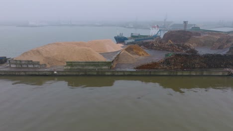 Aerial-establishing-view-of-wood-terminal-crane-loading-timber-into-the-cargo-ship,-Port-of-Liepaja-,-lumber-log-export,-overcast-day-with-fog-and-mist,-wood-chip-piles,-drone-moving-backward