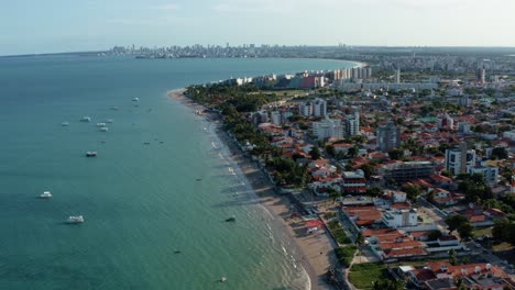 Tilt-up-aerial-drone-extreme-wide-shot-of-the-tropical-Bessa-beach-in-the-city-of-Joao-Pessoa,-Paraiba,-Brazil-with-people-enjoying-the-ocean,-tour-boats,-and-large-cityscape-in-the-background