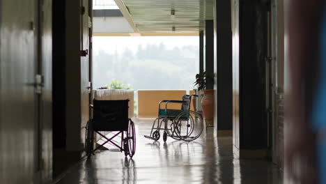 Zeitlupenaufnahme-Einer-Krankenschwester,-Die-Mit-Zwei-Leeren-Rollstühlen-Am-Ende-Des-Flurs-In-Einem-Ruandischen-Krankenhaus-Auf-Die-Kamera-Zuläuft