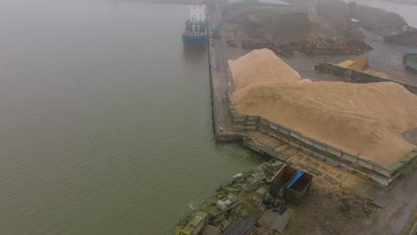 Aerial-establishing-view-of-wood-terminal-crane-loading-timber-into-the-cargo-ship,-Port-of-Liepaja-,-lumber-log-export,-overcast-day-with-fog-and-mist,-wide-drone-shot-moving-forward