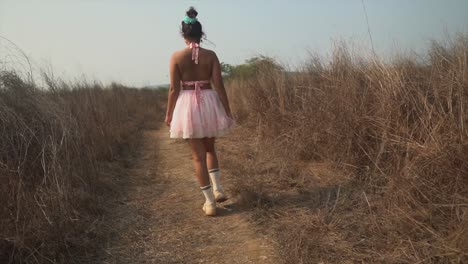 The-back-of-a-carefree-Asian-female-wearing-a-pink-tutu-dress-walking-along-a-remote-path-in-a-rural-grass-field-on-a-hot-summer’s-day-outdoors,-India