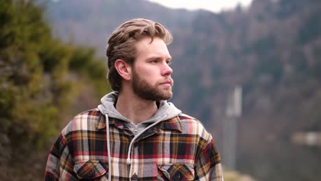 A-young-blond-man-in-a-checkered-hoodie-walks-beside-a-river,-his-tousled-hair-and-blue-eyes-catching-the-sunlight