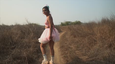 The-back-of-a-happy-carefree-Asian-female-walking-along-a-path-in-a-dry-grass-field-wearing-a-pink-tutu,-turning-around-playfully-holding-her-dress-and-smiling,-India