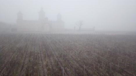 Aerial-forward-ascending-over-vineyard-with-Cos-d'Estournel-castle-in-background-on-foggy-day,-Bordeaux-in-France