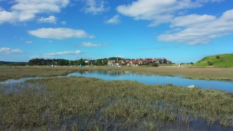 Low-Flying-drone-shot-across-marshes-revealing-reflection-in-the-water-of-blue-sky-and-fluffy-clouds-then-continues-towards-a-sailing-boat-and-the-village-of-Alnmouth,-Northumbria,-England