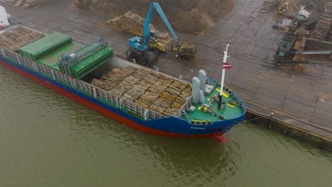 Aerial-establishing-view-of-wood-terminal-crane-loading-timber-into-the-cargo-ship,-Port-of-Liepaja-,-lumber-log-export,-overcast-day-with-fog-and-mist,-birdseye-drone-moving-backward