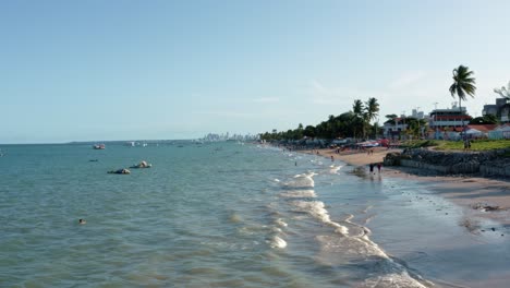 Dolly-in-aerial-extreme-wide-shot-of-the-tropical-Bessa-beach-in-the-capital-city-of-Joao-Pessoa,-Paraiba,-Brazil-with-people-enjoying-the-ocean,-small-fishings-boats-and-skyscrapers-in-the-background