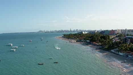 Truck-right-rising-aerial-drone-wide-shot-of-the-tropical-Bessa-beach-in-the-city-of-Joao-Pessoa,-Paraiba,-Brazil-with-people-enjoying-the-ocean,-tour-boats,-jet-ski,-and-skyscrapers-in-the-background
