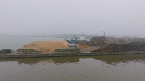 Aerial-establishing-view-of-wood-terminal-crane-loading-timber-into-the-cargo-ship,-Port-of-Liepaja-,-lumber-log-export,-day-with-fog-and-mist,-wood-chip-pile,-drone-shot-moving-forward