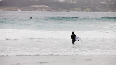 Weißer-Surfer-Im-Schwarzen-Neoprenanzug,-Der-Mit-Einem-Wachsbrett-Durch-Die-Wellen-Zum-Meer-Geht,-Um-In-Monterey-Bay,-Kalifornien,-Zu-Surfen