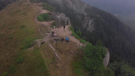 Aerial-helix-drone-shot-of-three-hikers-on-a-mountain-summit-near-a-steep-cliff