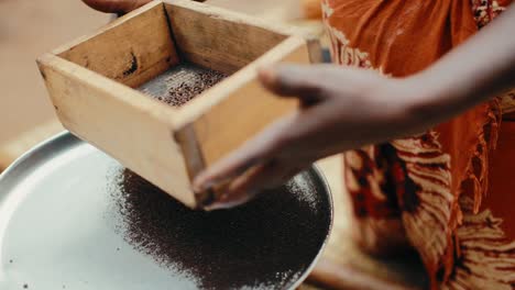 Black-Ugandan-lady-use-wooden-sifter-to-sieve-ground-coffee-onto-metal-plate