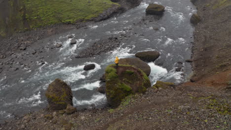 Aerial:-Panning-shot-of-a-man-wearing-yellow-jacket,-standing-on-a-rock-near-a-river
